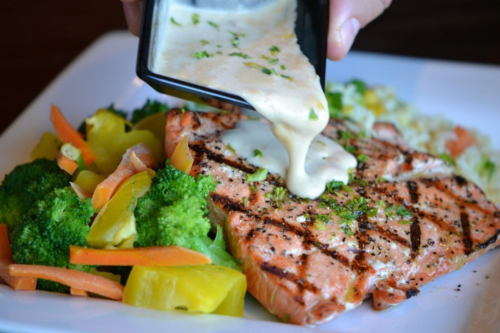 A plate of salmon and vegetables with a white sauce being ooured ofer it